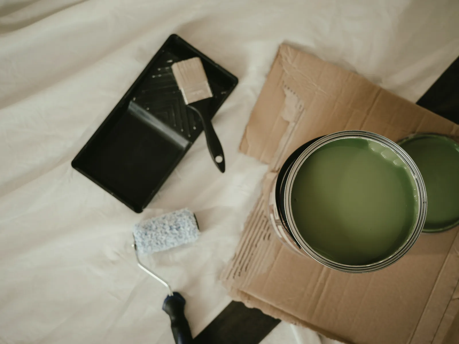 An open can of sage-green paint, a brush and a roller tray on a drop cloth