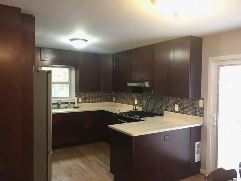 An opened-up kitchen with dark cherry cabinets, a glass mosaic backsplash, light counters and a wood floor